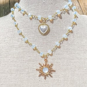 Puede incluir: Un collar de dos capas con un colgante de sol dorado y un colgante de corazón de perla. El collar está hecho de pequeñas cuentas transparentes y una cadena de oro.