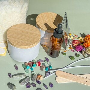 Pu&ograve; includere: Una vista dall'alto di accessori per la creazione di candele, tra cui due barattoli di candele bianche con coperchi in legno, una bottiglia di olio essenziale, fiori secchi, bastoncini di legno e pietre preziose colorate.