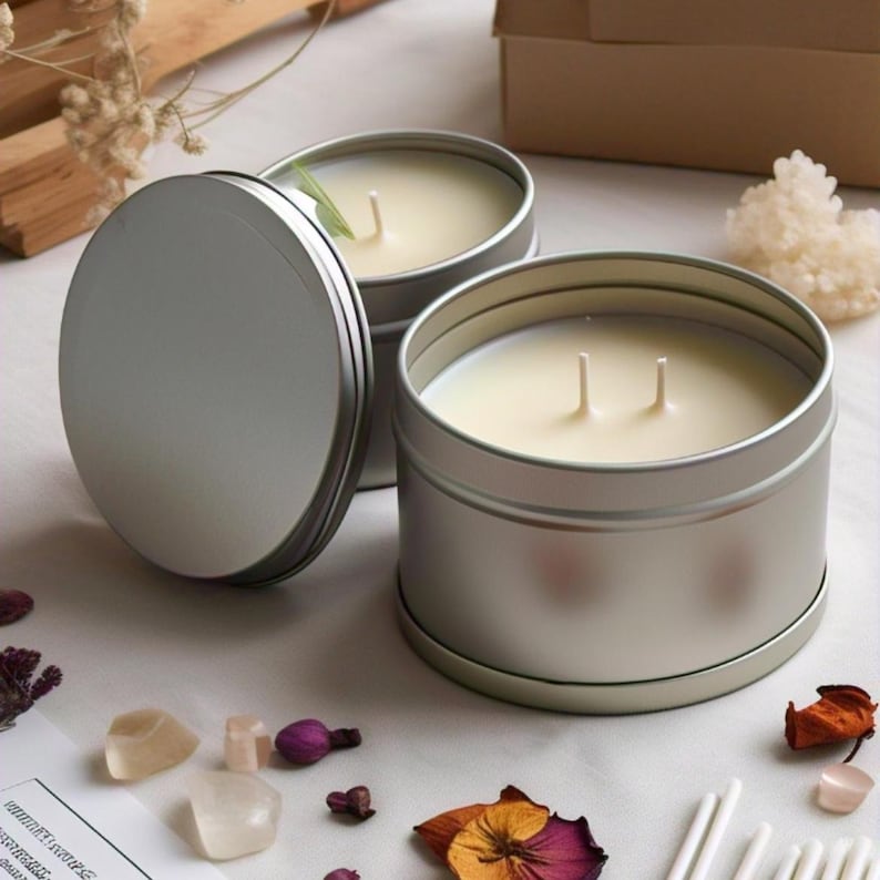 Pu&ograve; includere: Due barattoli di candela d'argento con coperchi. Le candele sono bianche e hanno stoppini. Le candele sono su una superficie bianca con fiori secchi e altri oggetti decorativi.