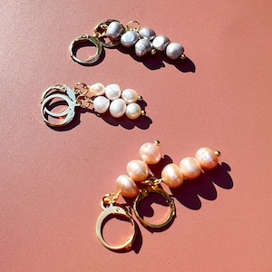 Puede incluir: Tres pares de pendientes de aro de oro con gotas de perlas. Los pendientes presentan perlas de diferentes tamaños y colores, incluyendo blanco, crema y gris.