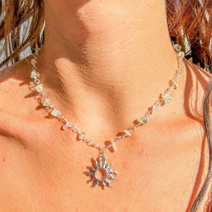 Puede incluir: Un collar de plata con un dije de sol y piedras transparentes. El collar está puesto en el cuello de una persona.
