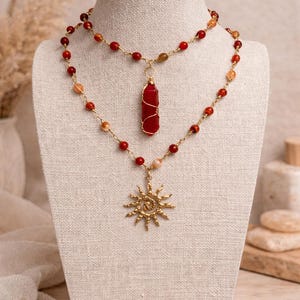 Puede incluir: Conjunto de dos collares expuestos en un maniquí. La hebra superior presenta cuentas rojas y naranjas con un colgante de cristal rojo. La hebra inferior tiene un colgante de sol dorado y cuentas similares. Los collares son dorados.