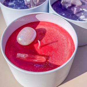 May include: A close-up of a red candle in a white container, with a white wick and two crystals. The candle has a smooth surface and is surrounded by two other candles with purple wax.