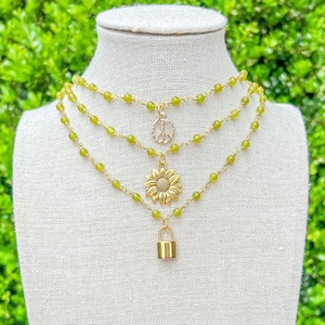 Puede incluir: Tres collares superpuestos con cuentas verdes y dijes dorados. Los dijes incluyen un signo de paz, un girasol y un candado.