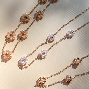 Puede incluir: Dos cadenas de oro con delicados abalorios con forma de flor. Una cadena presenta flores de perlas blancas y la otra flores de cristal marrón claro.