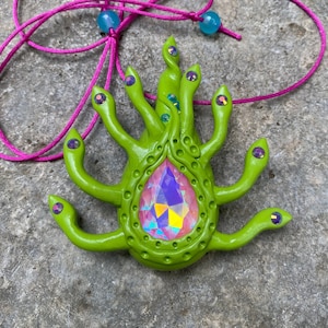 May include: A green, multi-armed pendant with a large iridescent gem in the center. The pendant is attached to a pink cord with blue beads.
