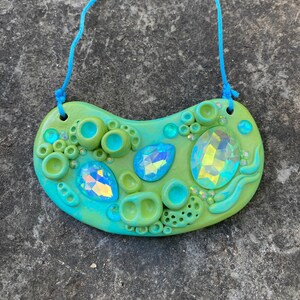 May include: A green and blue ceramic necklace with a whimsical design featuring iridescent gemstones, bubbles, and wavy lines. The necklace is strung on a blue cord.
