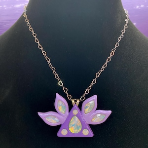 Puede incluir: Un collar con un colgante morado en forma de tri&aacute;ngulo adornado con una gran piedra preciosa iridiscente y piedras iridiscentes m&aacute;s peque&ntilde;as en cada punta del tri&aacute;ngulo. El colgante est&aacute; rodeado de cinco acentos en forma de flor morada.