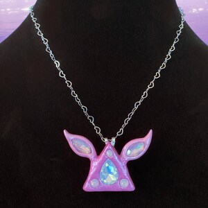 Puede incluir: Un collar con un colgante de tri&aacute;ngulo rosa adornado con cristales iridiscentes y una cadena en forma de coraz&oacute;n.