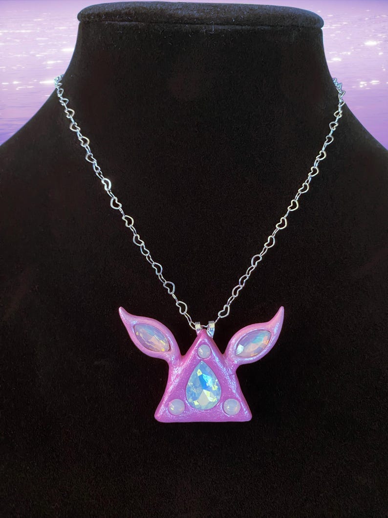 Puede incluir: Un collar con un colgante de tri&aacute;ngulo rosa adornado con cristales iridiscentes y una cadena en forma de coraz&oacute;n.