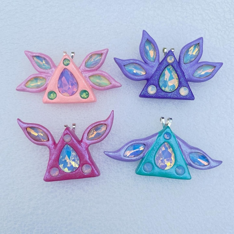 Puede incluir: Cuatro colgantes triangulares hechos a mano con alas iridiscentes. Cada colgante presenta una gran gema iridiscente en forma de l&aacute;grima en el centro y gemas iridiscentes m&aacute;s peque&ntilde;as alrededor de los bordes. Los colgantes son en tonos de rosa, morado, turquesa y verde.
