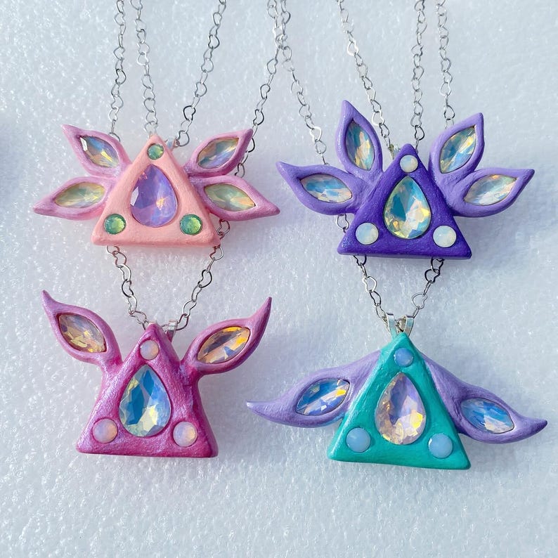 Puede incluir: Cuatro collares con colgantes triangulares con gemas iridiscentes y alas. Los colgantes son de color rosa, morado, verde y rosa oscuro. Las cadenas de los collares son de plata.