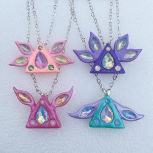 Puede incluir: Cuatro collares con colgantes triangulares con gemas iridiscentes y cadenas plateadas. Los colgantes son en tonos de rosa, morado, turquesa y rosa claro.