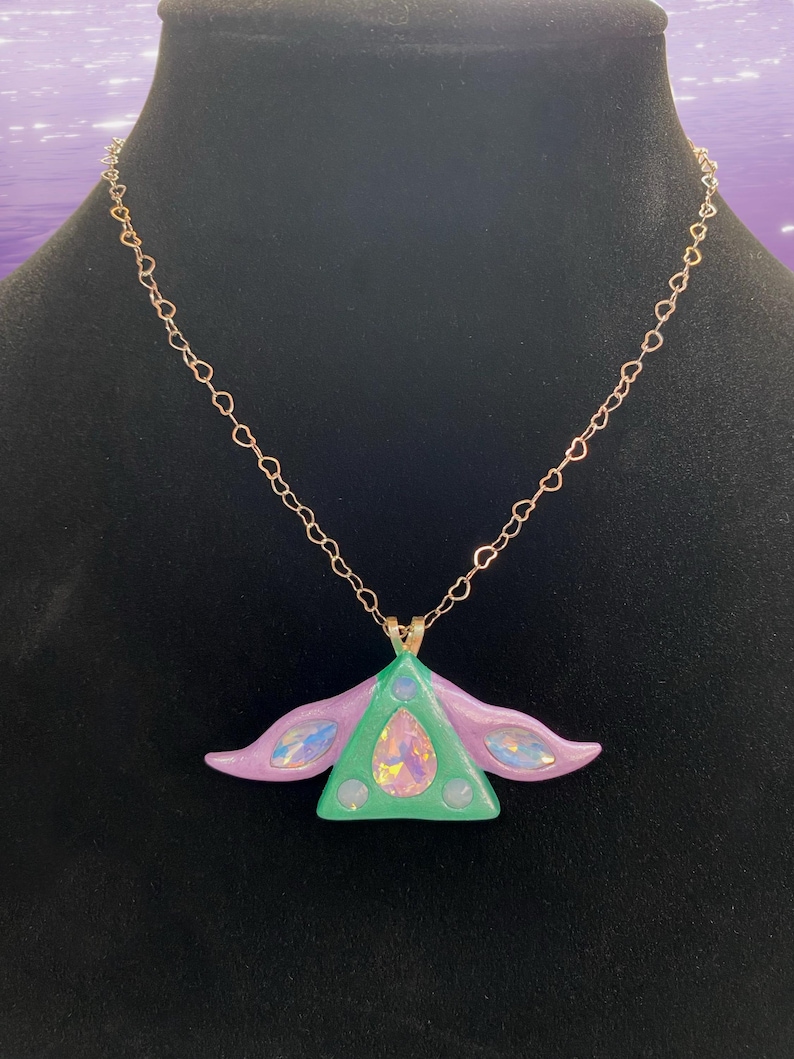 Puede incluir: Un collar con un colgante de tri&aacute;ngulo verde y morado con detalles iridiscentes que cuelga de una cadena de oro en forma de coraz&oacute;n.