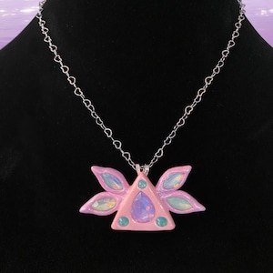Puede incluir: Un collar con un colgante de tri&aacute;ngulo rosa adornado con &oacute;palos iridiscentes y piedras verdes, rodeado de cinco alas rosas. El collar tiene una cadena plateada con eslabones en forma de coraz&oacute;n.