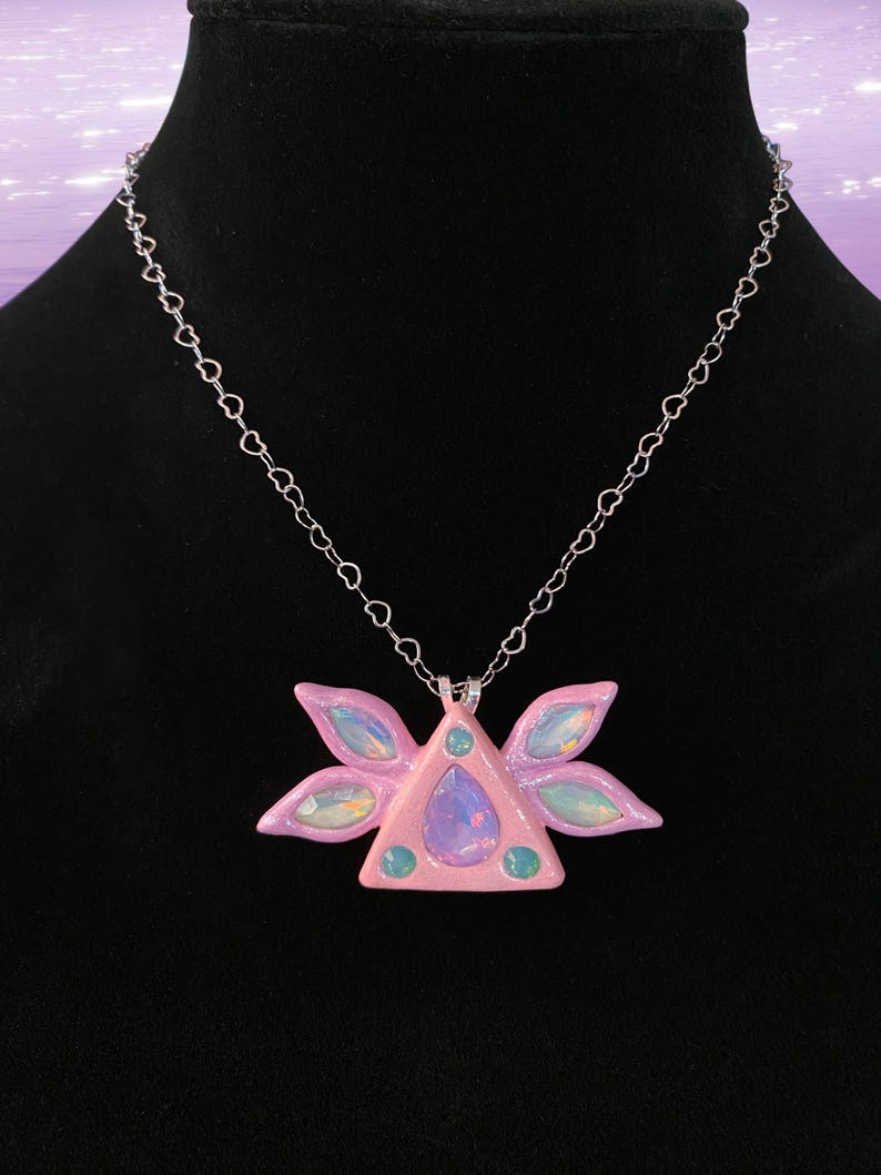 Puede incluir: Un collar con un colgante de tri&aacute;ngulo rosa adornado con &oacute;palos iridiscentes y piedras verdes, rodeado de cinco alas rosas. El collar tiene una cadena plateada con eslabones en forma de coraz&oacute;n.