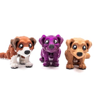 May include: Three small dog figurines with large, expressive eyes. The left figure is brown and white, the center figure is a glittery purple, and the right figure is a solid light brown. Each dog has floppy ears and a small black nose.