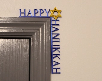 Könnte beinhalten: Eine blaue und goldene Hanukkah-Türkoration. Die Wörter "HAPPY" und "HANUKKAH" sind in blauen Buchstaben geschrieben. Ein goldener Davidstern befindet sich in der Ecke. Die Dekoration befindet sich auf einer grauen Tür und einer weißen Wand.