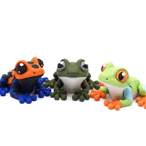 May include: Three colorful frog figurines. One frog is orange and black with blue legs, the second is green, and the third is green and orange. All have large, expressive eyes and are set against a white background.