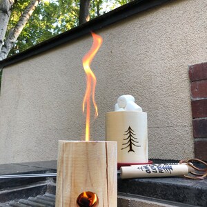 May include: A wooden fire pit with a flame burning inside. The fire pit is sitting on a black grill grate. There is a wooden cup with marshmallows next to the fire pit. The cup has a tree design carved into it. There is a wooden stick with the words "Campfire" and "Love" written on it.