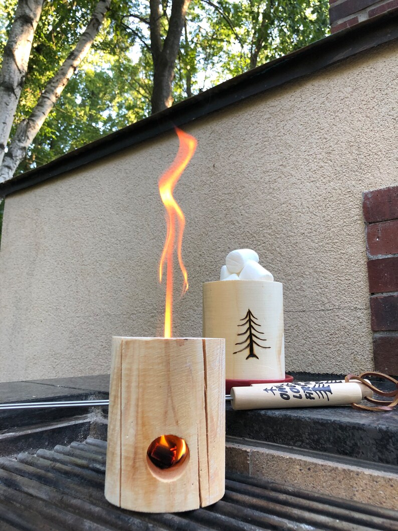 May include: A wooden fire pit with a flame burning inside. The fire pit is sitting on a black grill grate. There is a wooden cup with marshmallows next to the fire pit. The cup has a tree design carved into it. There is a wooden stick with the words "Campfire" and "Love" written on it.