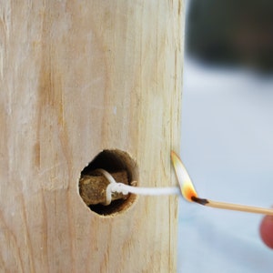May include: A white string is threaded through a hole in a wooden log. A lit match is being used to light the string.