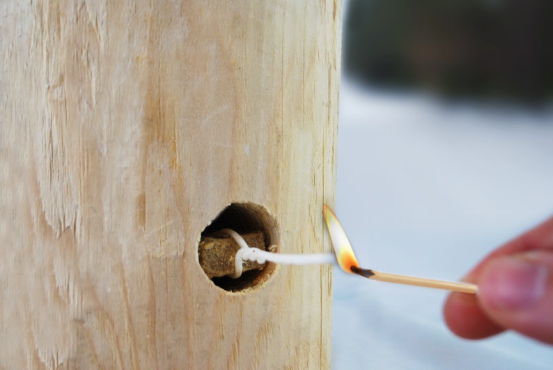 May include: A white string is threaded through a hole in a wooden log. A lit match is being used to light the string.