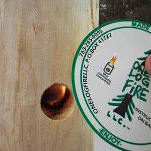 May include: A white round sticker with green text and a green pine tree design. The text reads "One Log Fire LLC. Made in Minnesota. 763-245-0095 P.O. Box 41722 S. MN 55441-0722 DNR Approved Firewood Instructions on Back -Enjoy- Peel Here".