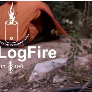 May include: A close-up of a OneLogFire log fire starter, a wooden log with a hole in the centre, burning with a bright orange flame. The log is surrounded by a metal grill and a tent is visible in the background.