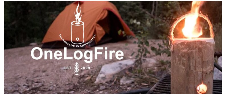 May include: A close-up of a OneLogFire log fire starter, a wooden log with a hole in the centre, burning with a bright orange flame. The log is surrounded by a metal grill and a tent is visible in the background.