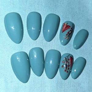 May include: A set of teal-colored, oval-shaped artificial nails. Two nails feature a design of a red leaf and berries with gold accents. The nails are arranged on a light blue surface.