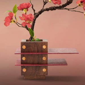 May include: A decorative floral arrangement featuring a bonsai-style tree with vibrant red flowers and green leaves. The tree is planted in a wooden cube-shaped planter, which sits atop a tiered display with red accents. The background is a gradient of pink.