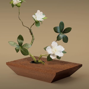 Puede incluir: Arreglo floral de ikebana de madera con flores blancas y hojas verdes. Las flores están dispuestas en un jarrón de madera geométrico marrón oscuro. El fondo es de color beige.
