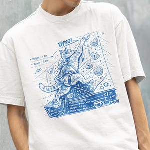 Könnte beinhalten: Weißes T-Shirt mit einem blauen Aufdruck einer Katze, die eine Wand erklimmt, mit dem Text "DYNO!". Das Design enthält Spezifikationen: Felis Catus, 4,5 kg Gewicht und MgCD-Kreide. Das Shirt hat einen Rundhalsausschnitt und kurze Ärmel.