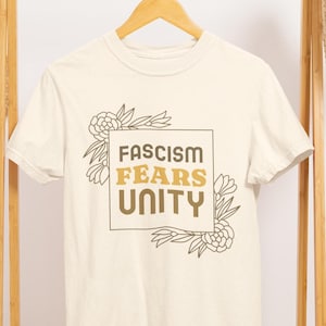 Może przedstawiać: Biała koszulka z brązowo-żółtym nadrukiem z napisem "Fascism Fears Unity" otoczonym wzorami kwiatowymi.