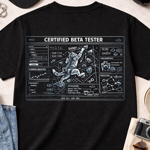 May include: Black t-shirt with a graphic featuring a climbing cat and the text "CERTIFIED BETA TESTER." The design includes climbing analytics, paw biometrics, and a route plan. The cat is climbing a wall with the text "DYNO!" above it.