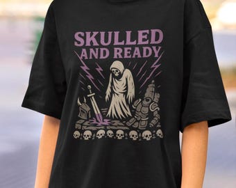 Pode incluir: Camiseta preta com um design gráfico apresentando uma figura encapuzada, uma espada e uma armadura, com o texto "SKULLED AND READY" em roxo. O design inclui raios e uma fileira de caveiras.