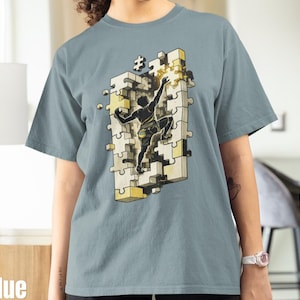 Könnte beinhalten: Eisblaues T-Shirt mit einer Grafik einer Person, die ein Puzzle erklimmt. Das Puzzle besteht aus weißen und gelben Teilen. Die Person ist eine schwarze Silhouette, die nach einem Puzzleteil greift. Das Shirt ist ein bequemer, lässiger Stil.
