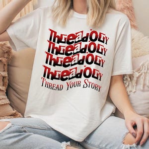 Pode incluir: Uma camiseta branca com o texto "Thread Your Story" em um design empilhado em vermelho e preto. A camiseta está sendo usada por uma pessoa sentada, usando jeans azul claro.