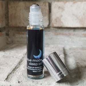 Könnte beinhalten: Eine blaue Glas-Rollflasche mit silbernem Verschluss und einem Design mit einem blauen Mond. Das Etikett lautet "blue moon sleep oil" und "blend of essential oils, crystals & herbs". Die Flasche enthält eine klare Flüssigkeit mit Glitzer und hat ein Fassungsvermögen von 10 ml / 0,34 oz.