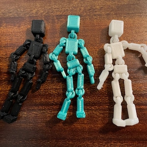 May include: Three articulated figures in black, teal, and white, each with a block-shaped head and jointed limbs. The figures are posed on a wooden surface, showcasing their flexibility and design. These are likely used for art or animation.