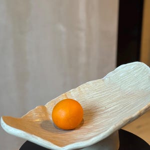 Könnte beinhalten: Eine beige Keramikservierschale mit strukturierter Oberfläche, die eine einzelne orangefarbene Frucht enthält. Die Schale hat eine organische Form und steht auf einem kleinen, runden Sockel. Die Frucht hat eine leuchtend orange Farbe.