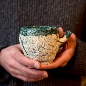 Könnte beinhalten: Handgefertigte Keramik-Tasse mit einem strukturierten, cremefarbenen Körper und einem dunkelgrünen Rand und Henkel. Die Tasse hat ein rustikales, handwerkliches Aussehen, perfekt für den Genuss von Kaffee oder Tee. Die Tasse wird mit beiden Händen gehalten.