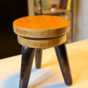 Op de afbeelding: Een kleine houten kruk met een ronde zitting en drie donkerbruine, taps toelopende poten. De zitting is lichtbruin en het middengedeelte heeft een lichtere houttint. Een eenvoudig, functioneel meubelstuk.