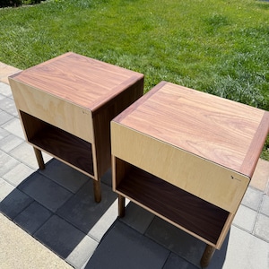 May include: Two square bedside tables featuring a dark brown wood top and a light beige wood front panel. Each table has an open storage compartment with a dark brown interior and is supported by four cylindrical wooden legs.