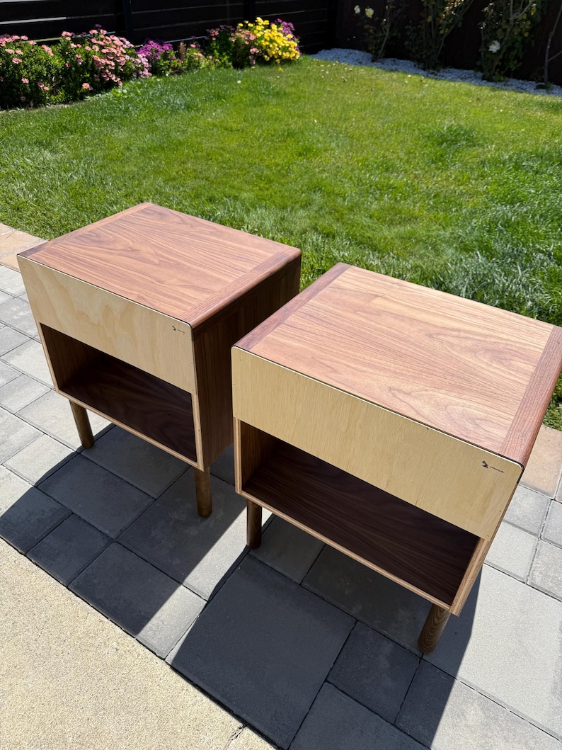 May include: Two square bedside tables featuring a dark brown wood top and a light beige wood front panel. Each table has an open storage compartment with a dark brown interior and is supported by four cylindrical wooden legs.