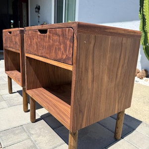 May include: Two matching dark brown wooden nightstands featuring a single drawer with a recessed handle and an open storage shelf below. Each piece is supported by four cylindrical legs and displays a prominent wood grain pattern throughout the surface.