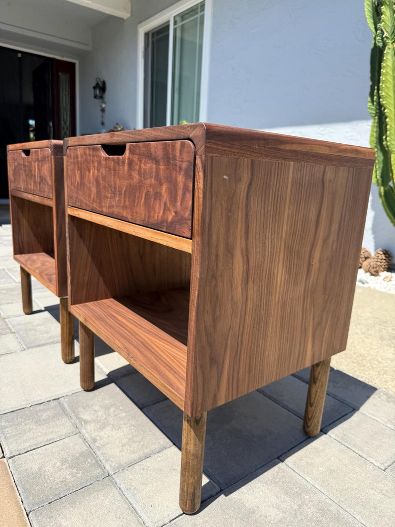 May include: Two matching dark brown wooden nightstands featuring a single drawer with a recessed handle and an open storage shelf below. Each piece is supported by four cylindrical legs and displays a prominent wood grain pattern throughout the surface.
