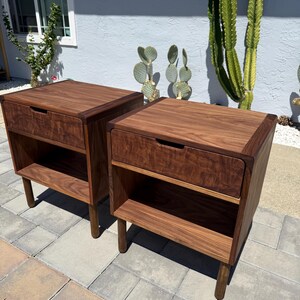 May include: A pair of matching dark brown wooden nightstands, each featuring a single top drawer with a recessed handle and an open storage shelf below. The nightstands are supported by four cylindrical legs and display a prominent wood grain.
