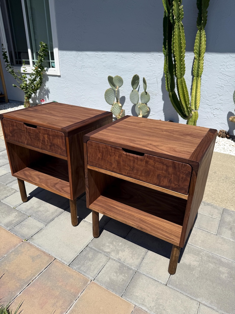 May include: A pair of matching dark brown wooden nightstands, each featuring a single top drawer with a recessed handle and an open storage shelf below. The nightstands are supported by four cylindrical legs and display a prominent wood grain.